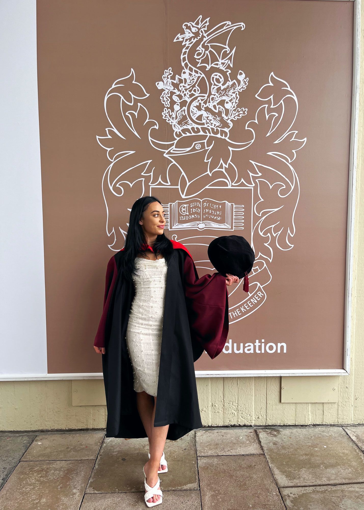 Liv stood in her Essex graduation robes holding a PhD cap in her left hand. She is stood in front of the Essex crest in gold