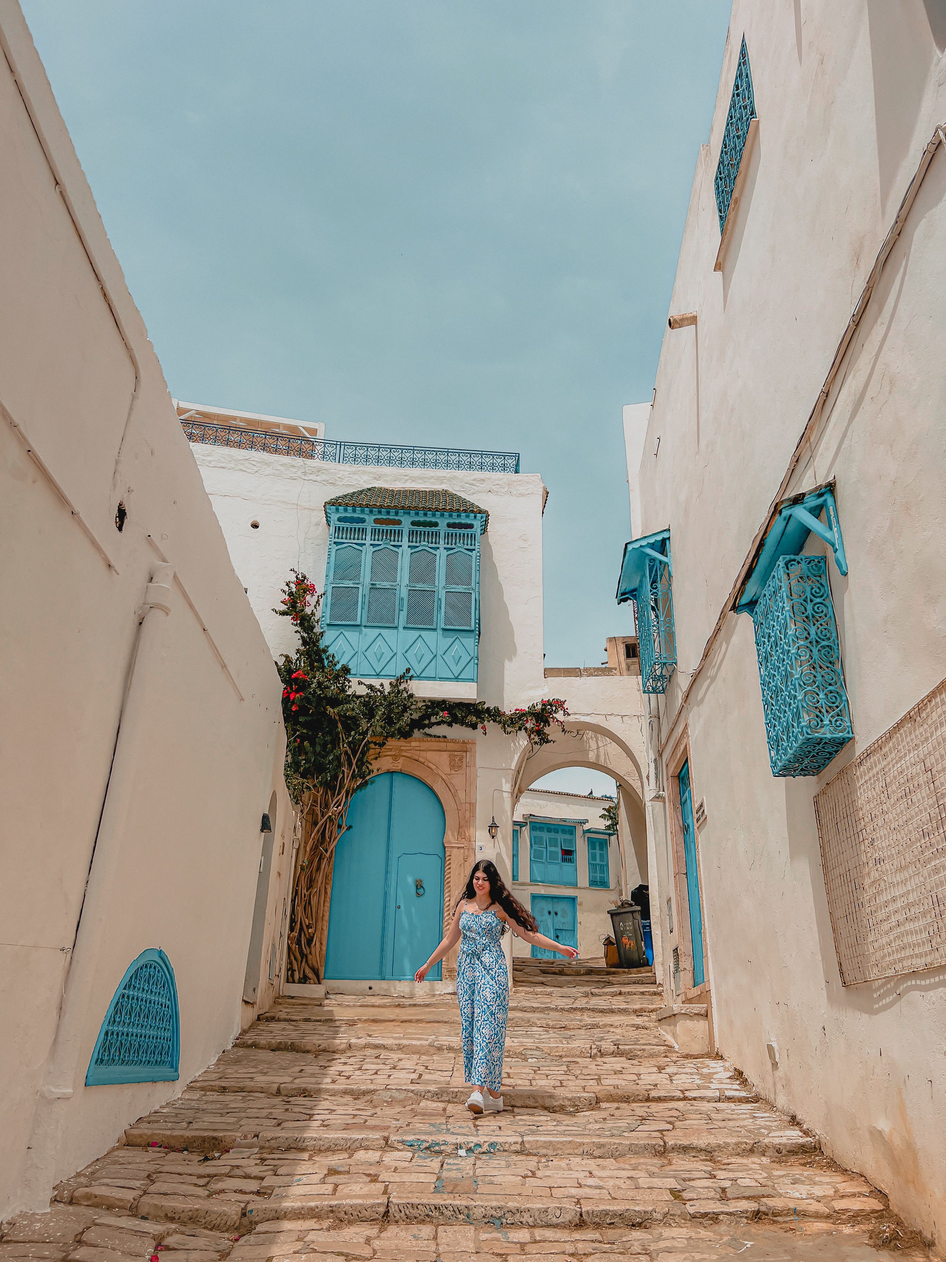Ela is walking towards the camera, the backdrop is an old Tunisian street with soft brown paintwork and a bright blue doorway. There are flowers and greenery around a door.