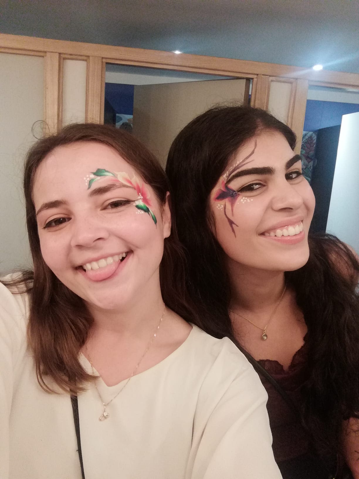 Two female friends are smiling at the camera, it's a close up shot of their faces. They both have glitter/face paint around their eyes