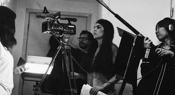 A black and white image of Ela stood using a video camera, there are other crew members around her holding boom mics and lights