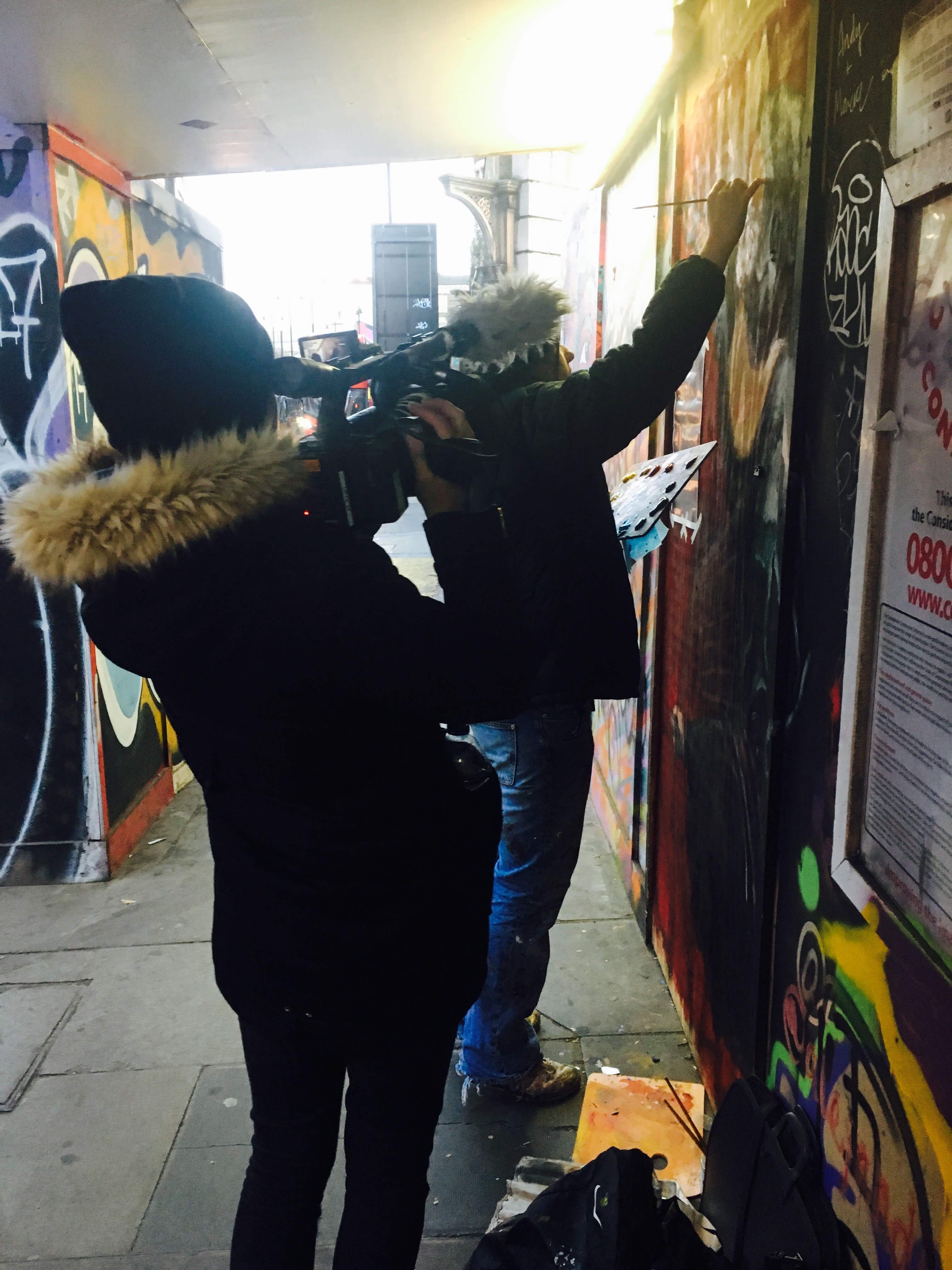 Ela is holding a camera, filming a street artist while they paint a wall