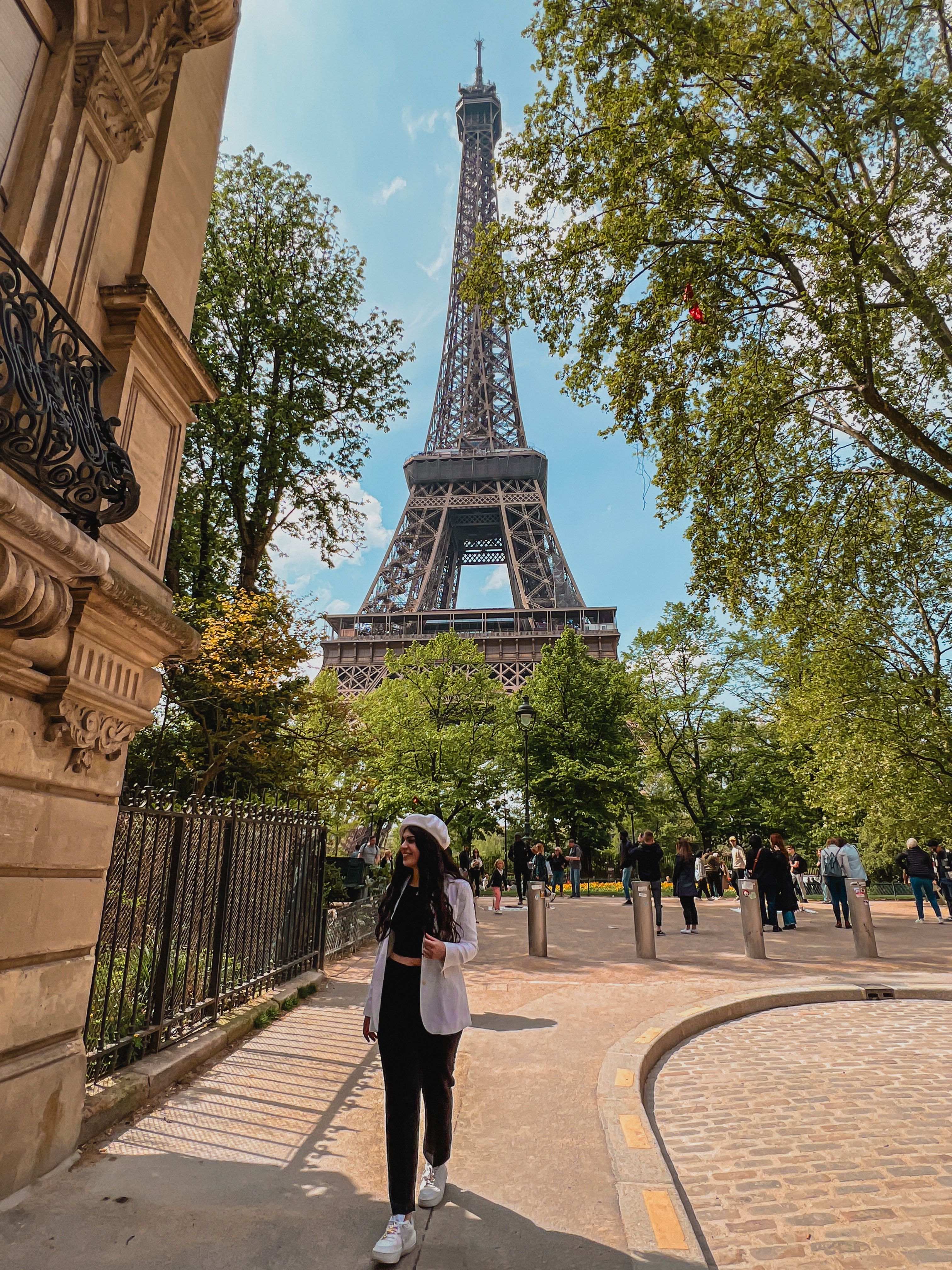 Ela is walking towards the camera, smiling. In the background you can see the Eiffel tower. The sky is blue and the trees in the background have soft green leaves.