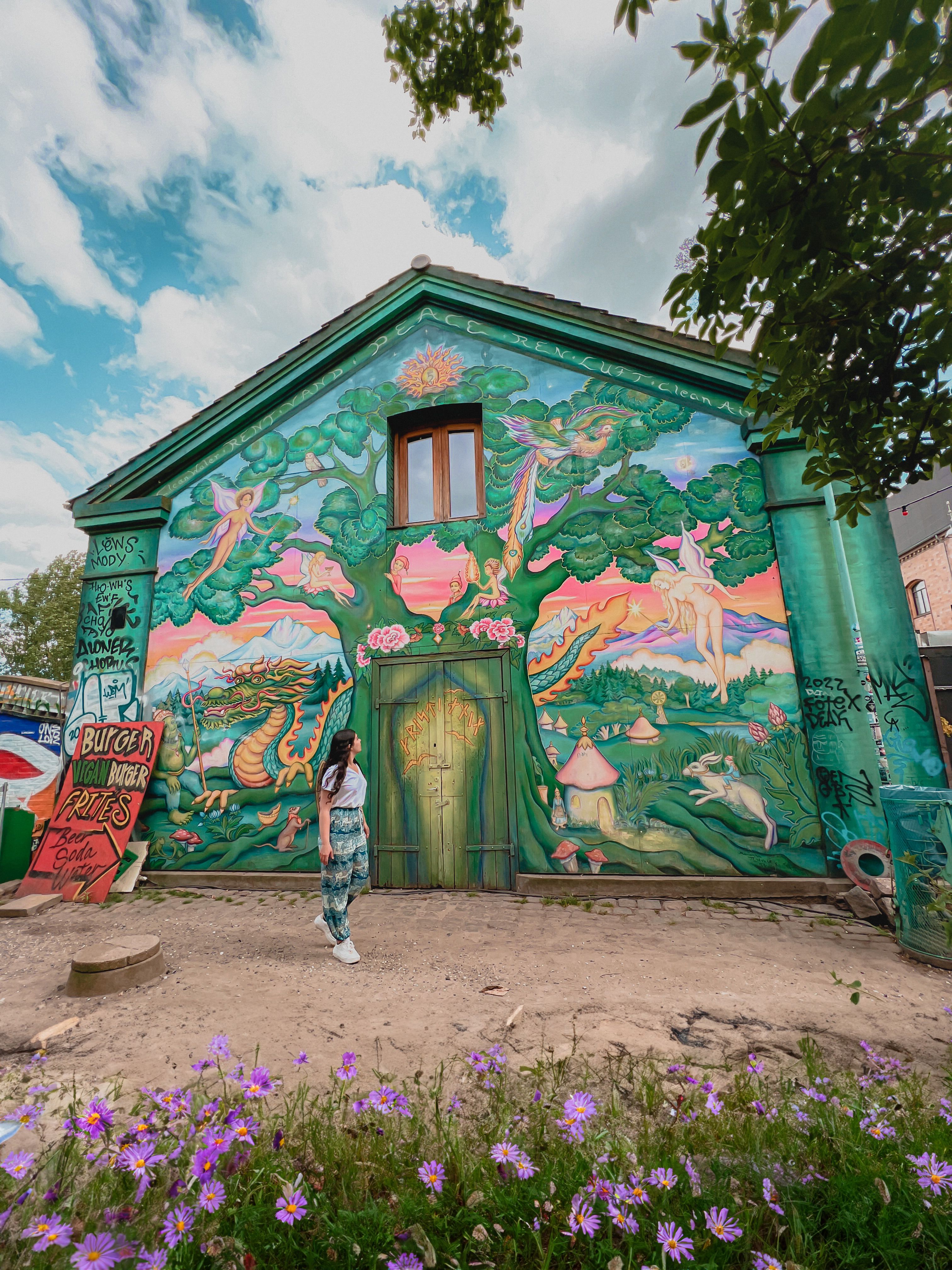 Ela walking by a large mural on the side of a building. The mural is of a large green tree with fairies, a dragon and other mythical creatures around it