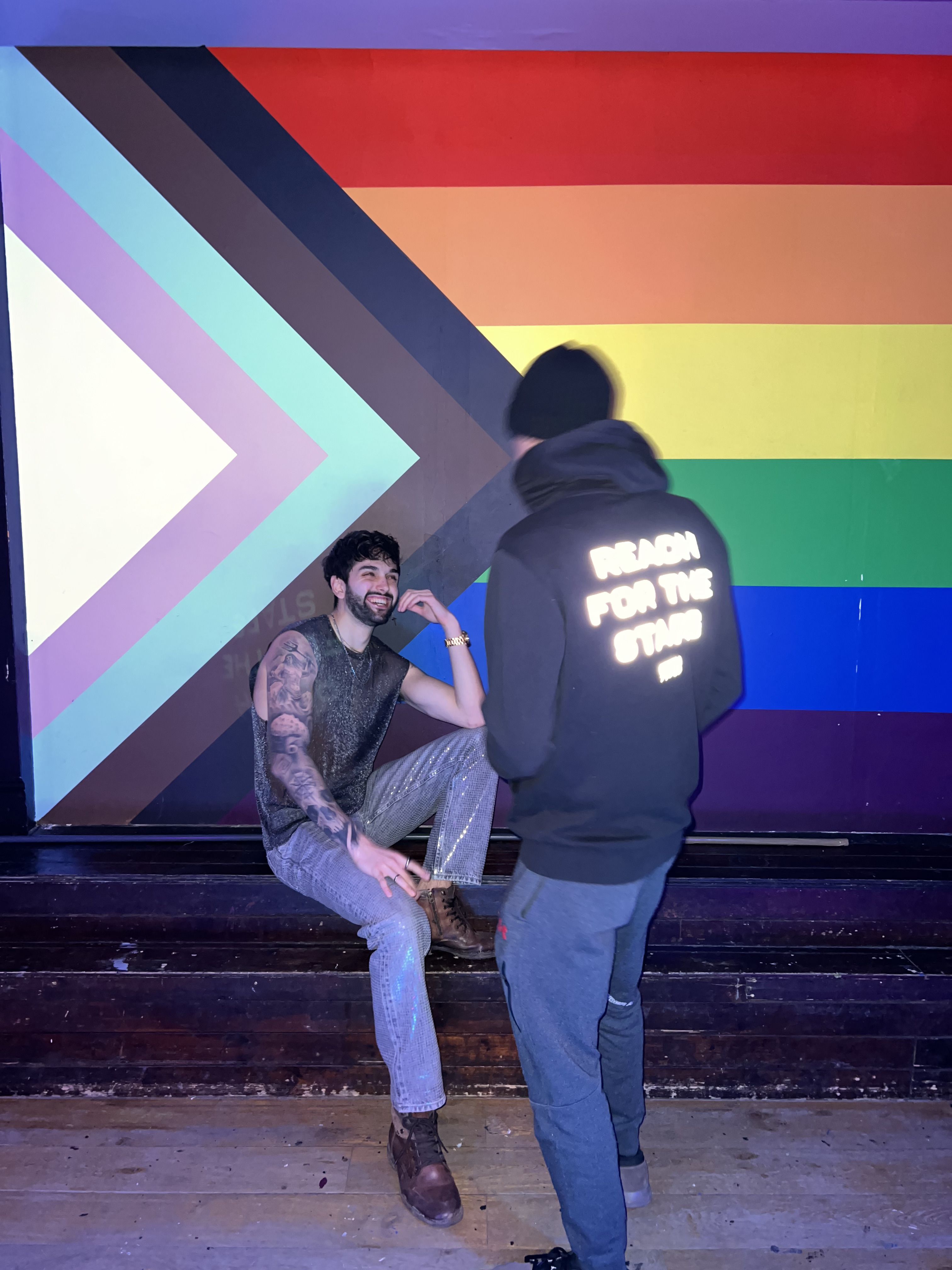 Michael sat against a rainbow wall laughing with a friend whose hoodie has "reach for the stars" written on the back